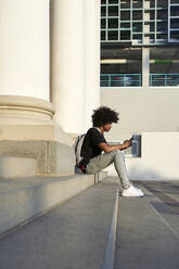 Junger Mann hört Musik und hält sein Smartphone, sitzt auf einer Treppe - VEGF01768