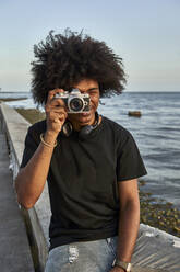 Young man sitting on wall and photographing with his camera at seafront - VEGF01774