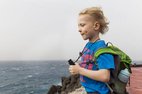 Junge mit Fernglas und Rucksack schaut auf das Meer, während er an der Costa Adeje, Kanarische Inseln, Spanien, steht - IHF00283