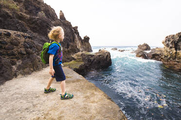 Junge in voller Länge, der auf einer Felsformation steht, während er das Meer an der Costa Adeje, Kanarische Inseln, Spanien, erkundet - IHF00285