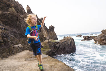 Fröhlicher Junge, der auf einer Felsformation am Meer läuft, während er seinen Urlaub an der Costa Adeje, Kanarische Inseln, Spanien, genießt - IHF00286