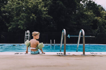 Rear View Of Shirtless Boy Wearing Water Wings Sitting At Poolside - EYF01084
