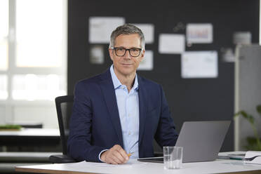 Portrait of businessman with laptop at desk in office - RBF07157