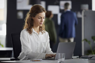 Geschäftsfrau mit Laptop am Schreibtisch im Büro mit Kollegen im Hintergrund - RBF07172