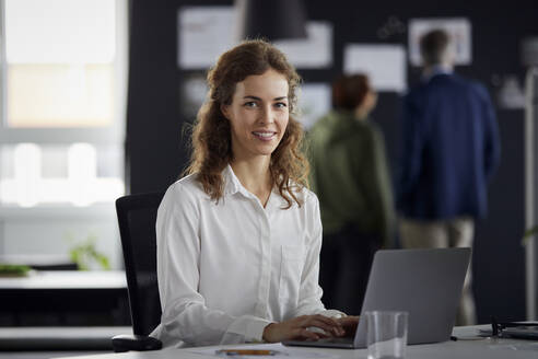 Porträt einer lächelnden Geschäftsfrau mit Laptop am Schreibtisch im Büro mit Kollegen im Hintergrund - RBF07173