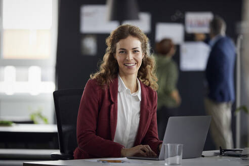 Porträt einer lächelnden Geschäftsfrau mit Laptop am Schreibtisch im Büro mit Kollegen im Hintergrund - RBF07175