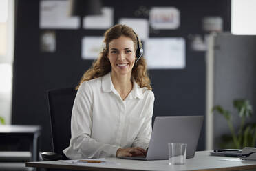 Portrait of smiling businesswoman with headset and laptop at desk in office - RBF07177