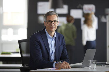 Portrait of businessman at desk in office with colleagues in background - RBF07178