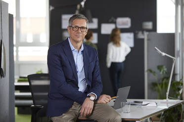 Portrait of businessman on desk in office with colleagues in background - RBF07180