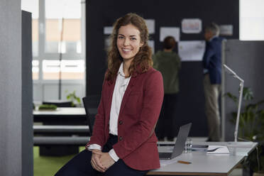 Portrait of smiling businesswoman sitting on desk in office with colleagues in background - RBF07185