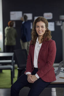 Porträt einer lächelnden Geschäftsfrau, die auf einem Schreibtisch im Büro sitzt, mit Kollegen im Hintergrund - RBF07186