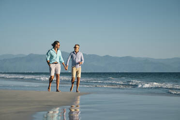 Reife schwule Männer in voller Länge, die sich an den Händen halten, während sie am Strand gegen den klaren blauen Himmel spazieren gehen, Riviera Nayarit, Mexiko - ABAF02293