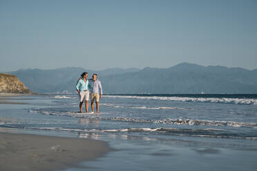 Ältere schwule Männer halten sich an den Händen, während sie am Strand gegen den klaren blauen Himmel spazieren gehen, Riviera Nayarit, Mexiko - ABAF02295