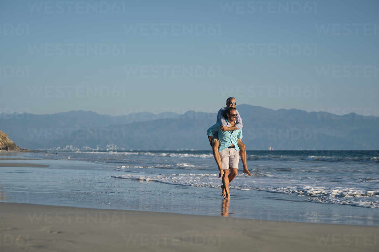 Mature gay man giving piggyback to partner while walking on shore at beach  against clear blue