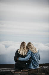 Rückansicht von Freundinnen mit Blick auf eine idyllische Wolkenlandschaft beim Sitzen auf dem Vulkan Haleakala, Hawaii, Maui, USA - LHPF01198