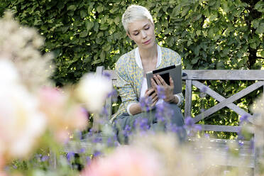 Porträt einer Frau, die sich auf einer Holzbank in einem Park ausruht und auf ein digitales Tablet schaut - FLLF00430