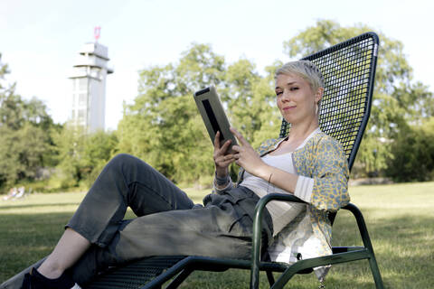 Porträt einer lächelnden Frau mit digitalem Tablet, die auf einem Liegestuhl in einem Park sitzt, lizenzfreies Stockfoto