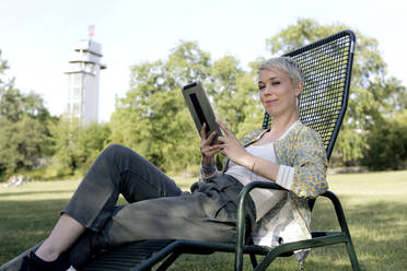 Porträt einer lächelnden Frau mit digitalem Tablet, die auf einem Liegestuhl in einem Park sitzt - FLLF00432