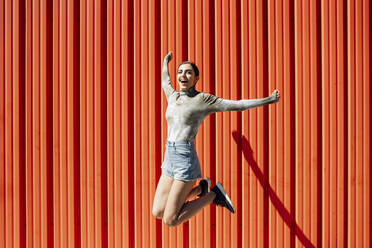 Full length of carefree young woman jumping against red corrugated wall on sunny day - JSMF01475
