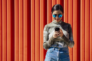 Lächelnde junge Frau, die ein Smartphone benutzt, während sie an einer roten Wellblechwand an einem sonnigen Tag steht - JSMF01478