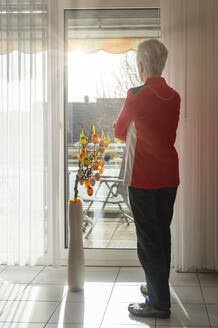 Senior woman standing at closed balcony and looking out of the window - FRF00907
