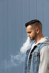 Junger Mann raucht mit einer elektronischen Zigarette auf blauem Hintergrund - CAVF77185