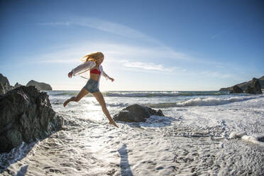 Teenage girl jumping off rock into surf at beach - CAVF77240
