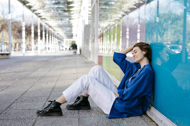 Junge Frau mit urbanem Look sitzt auf dem Boden und lehnt sich an eine bunte Glaswand mit ihrem Spiegelbild - TCEF00264