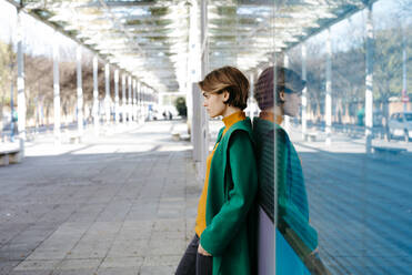 Porträt einer jungen Frau mit grünem Mantel, die sich an eine bunte Glaswand mit ihrem Spiegelbild lehnt - TCEF00266
