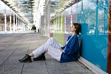 Junge Frau mit urbanem Look sitzt auf dem Boden und lehnt sich an eine bunte Glaswand mit ihrem Spiegelbild - TCEF00276