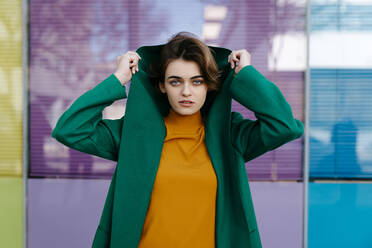 Young woman wearing putting on hood of her green coat in front of purple glass pane - TCEF00286