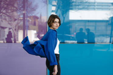 Porträt einer jungen Frau, die mit der Bewegung ihrer Jacke vor einer bunten Glaswand spielt - TCEF00297