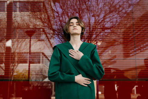 Young woman with closed eyes wearing green coat in front of a glass pane - TCEF00300