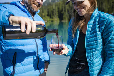 Mann schenkt seiner Partnerin am See ein Glas Wein ein. - CAVF77464