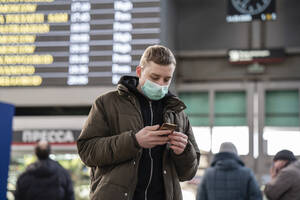 Young man with face mask at train station in the city, using smartphone - VPIF02127