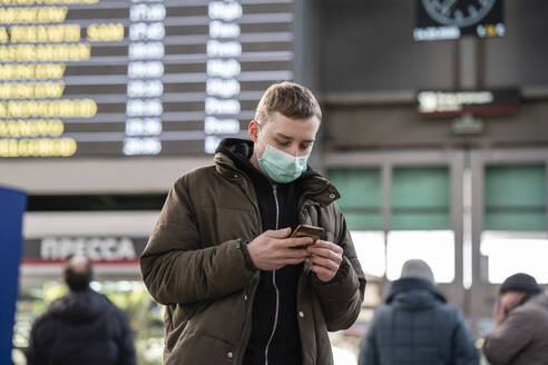 Junger Mann mit Gesichtsmaske am Bahnhof in der Stadt, der sein Smartphone benutzt - VPIF02127