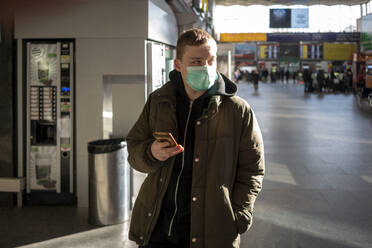 Young man with face mask at train station in the city, holding smartphone - VPIF02128