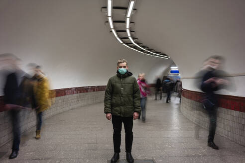 Young man with face mask standing isolated in subway underpass, with people moving around him - VPIF02129