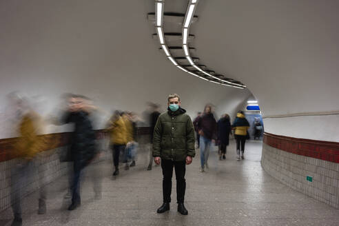 Young man with face mask standing isolated in subway underpass, with people moving around him - VPIF02130