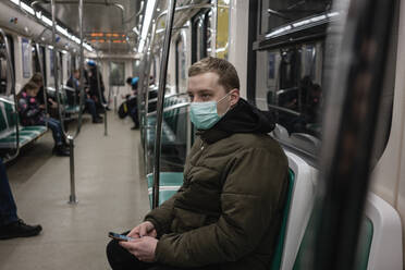 Junger Mann in einem Nahverkehrszug, mit Gesichtsmaske, der ein Smartphone benutzt - VPIF02133