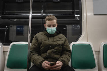 Junger Mann in einem Nahverkehrszug, mit Gesichtsmaske, der ein Smartphone benutzt - VPIF02134