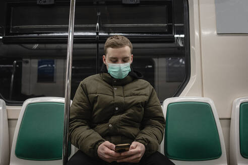 Young man in commuter train, wearing face mask, using smartphone - VPIF02134