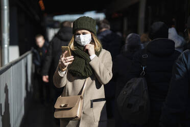 Frau mit Gesichtsmaske steht an einer Bushaltestelle und benutzt ein Smartphone - VPIF02137
