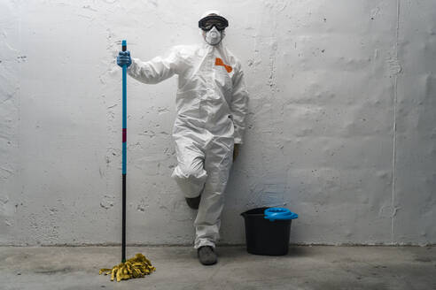 Woman with cleaning equipment, wearing protective clothing, leaning against wall - DLTSF00651