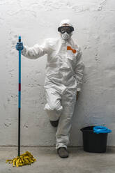Woman with cleaning equipment, wearing protective clothing, leaning against wall - DLTSF00652
