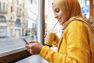 Porträt einer glücklichen jungen Frau mit Smoothie und Smartphone in einem Café - WPEF02716