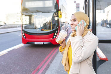 Porträt einer lächelnden jungen Frau am Telefon, die an der Bushaltestelle wartet - WPEF02719