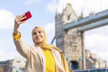 Porträt einer lächelnden jungen Frau, die ein Selfie mit ihrem Smartphone vor der Tower Bridge macht, London, UK - WPEF02721