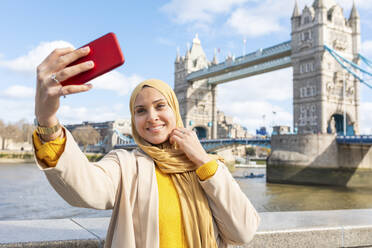 Porträt einer lächelnden jungen Frau, die ein Selfie mit ihrem Smartphone vor der Tower Bridge macht, London, UK - WPEF02722