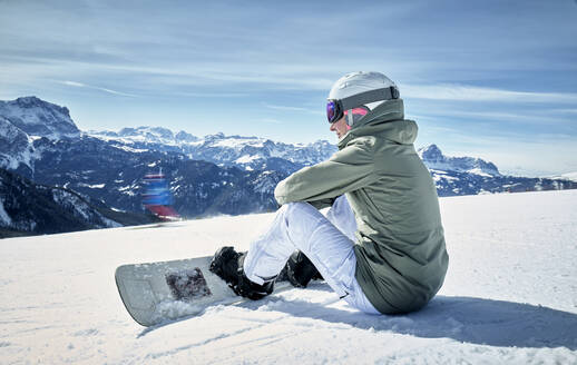 Lächelnde Frau sitzt mit Snowboard auf einem Aussichtspunkt und genießt die Aussicht in der Ferne - DIKF00391